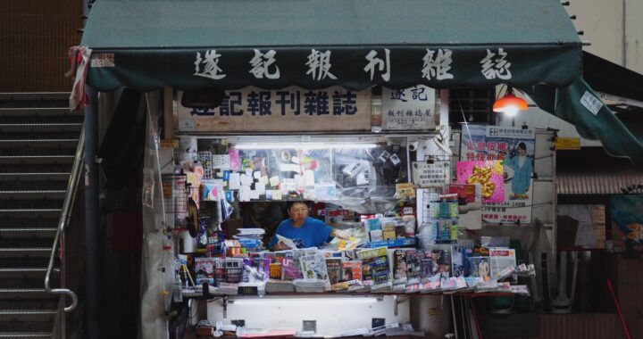 Fotografia em cores de banca de jornal em Hong Kong. No centro, dentro da banca, a dona do estabelecimento.