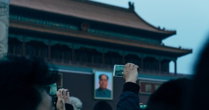 Fotografia em cores da Porta da Paz Celestial. Em primeiro plano, uma mão ergue um celular. Ao fundo vê-se o retrato de Mao.