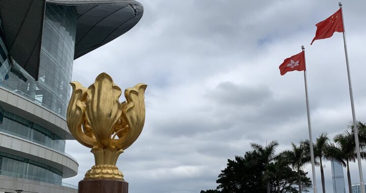 Fotografia em cores do Golden Bauhinia Square, onde ocorrem celebrações da transferência de soberania de Hong Kong. Vê-se a flor-símbolo de Hong Kong, uma escultura dourada, à frente; ao fundo, as bandeiras da China e de Hong Kong.
