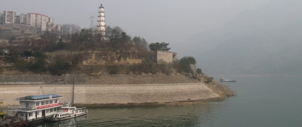 Fotografia em cores do rio Yangtze. Vê-se um barco na água e um trecho da margem se elevando, com prédios chineses tradicionais e modernos ao fundo. Há uma névoa ofuscando o fundo da paisagem.