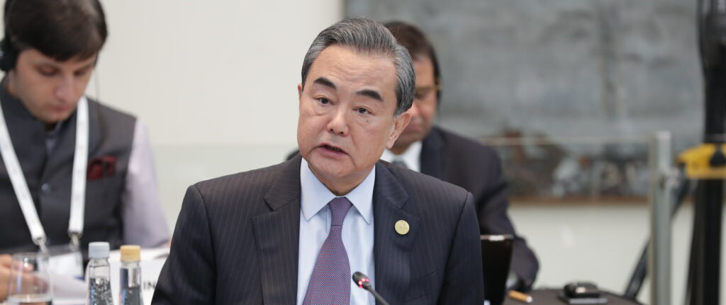 Wang Yi, ministro das Relações Exteriores da China vestindo um terno preto com risca, camisa azul clara e gravata roxa sentado em mesa em frente a placa China