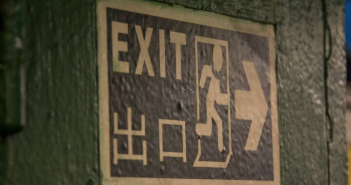 Fotografia em cores de placa verde e branca indicando saída em inglês e chinês.
