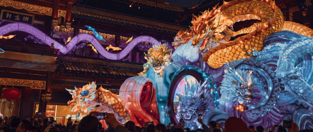 Fotografia em cores de apresentação em templo chinês assistida por um grupo de pessoas. Uma enorme lanterna de papel representa diferentes dragões chineses.
