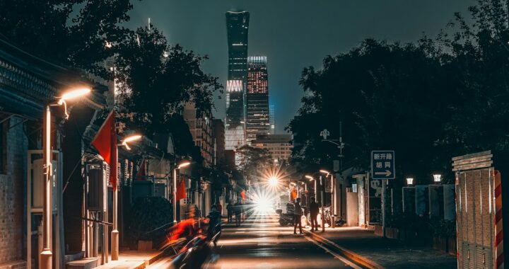 Fotografia em cores de rua de Pequim à noite, com calçadas iluminadas e arranha-céus ao fundo.